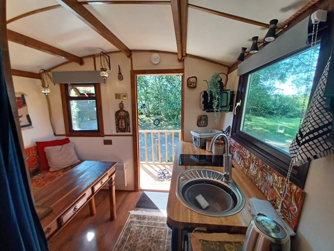 A welcoming interior features a cozy sitting area with a cushioned bench and decorative accents. Natural light filters through a window, illuminating the kitchenette equipped with a sink and wooden counter. A doorway leads to an outdoor area, framed by lush greenery.