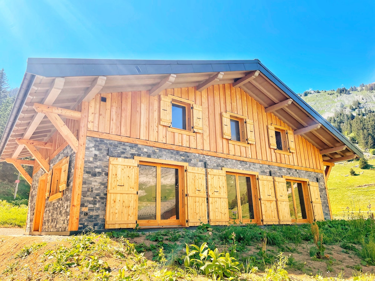A charming chalet showcases a combination of wooden and stone exterior elements. Large windows with wooden shutters allow natural light to enter, while the surrounding landscape features green hills under a clear blue sky.