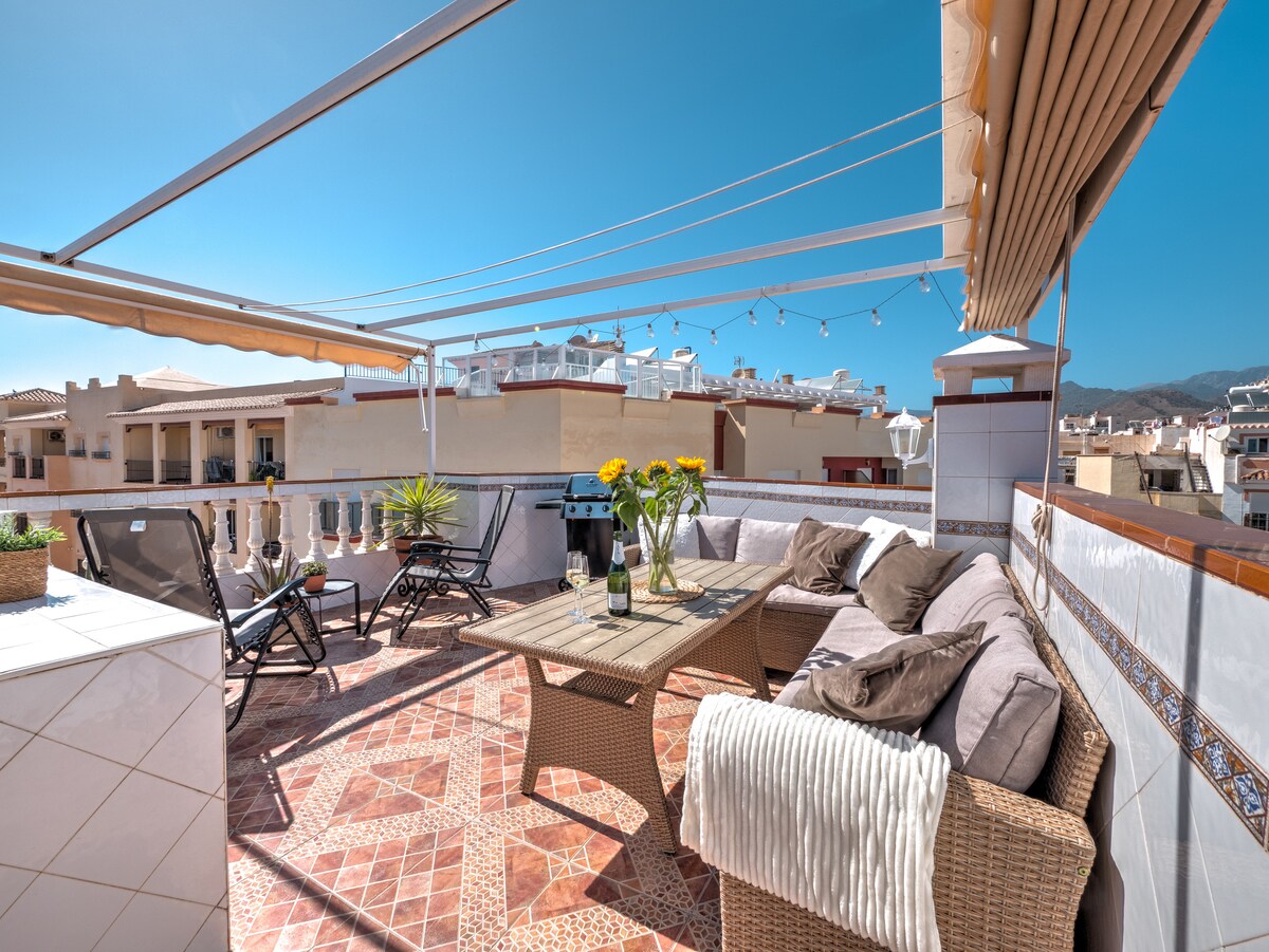 The spacious private rooftop terrace is furnished with a comfortable seating area and a dining table. A BBQ grill is visible, surrounded by potted plants. Loungers invite relaxation while a retractable awning provides shade, enhancing the panoramic views of the surrounding area.