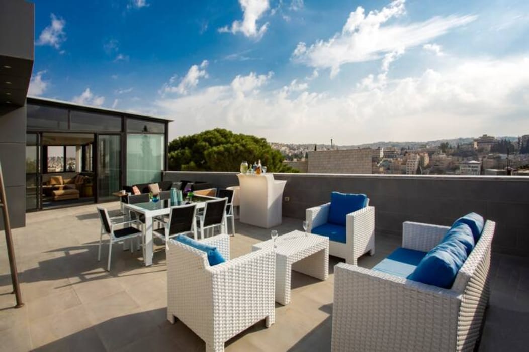 An outdoor terrace offers a comfortable seating area with white wicker furniture accented by blue cushions. A dining table set for six is positioned nearby, alongside a bar area. The backdrop features panoramic views of the surrounding city under a partly cloudy sky.