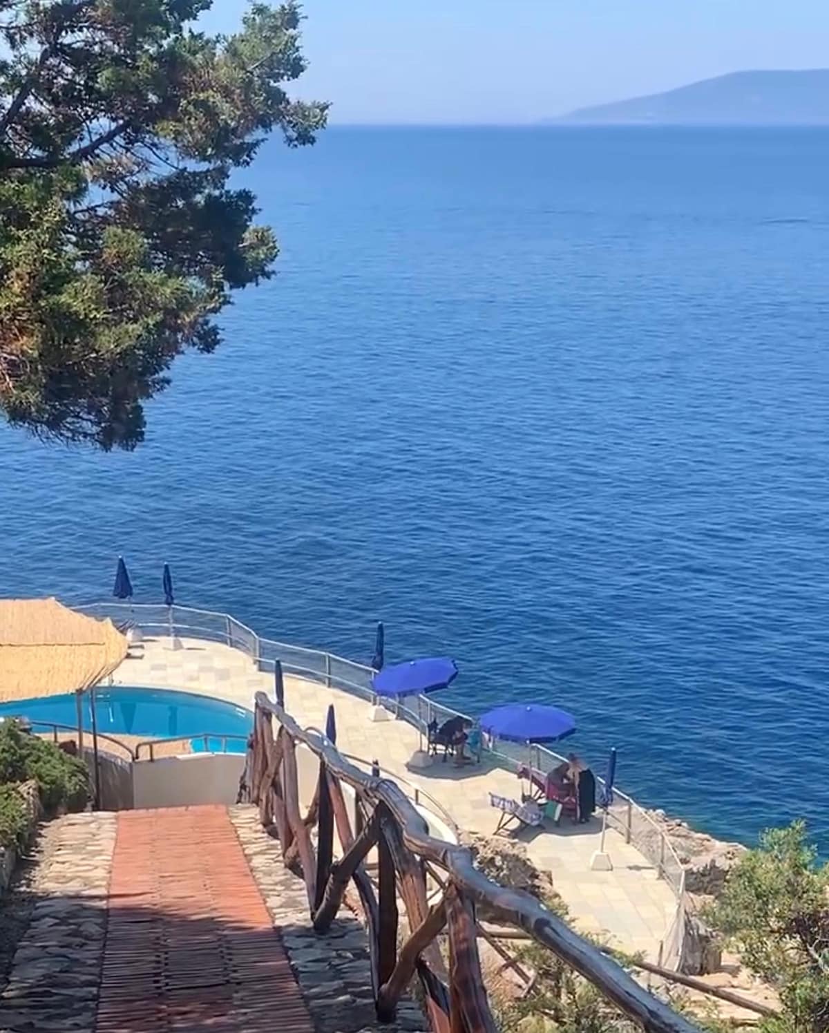 A serene pool area overlooks clear blue waters, surrounded by vibrant umbrellas. Stone pathways lead down from a scenic viewpoint. The spacious deck features lounge chairs, offering a perfect spot for relaxation while enjoying the tranquil marine environment.