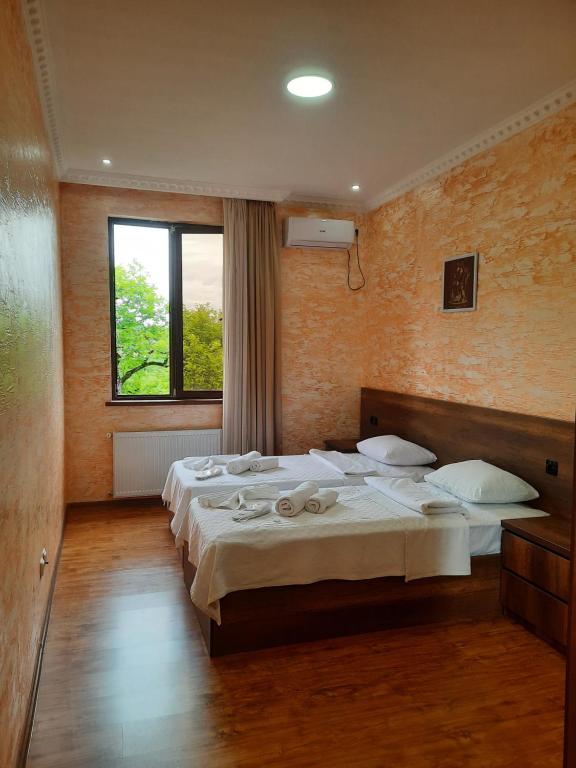 A guest room with warm orange walls features two spacious beds with neatly arranged towels. Natural light enters through a large window draped with light curtains, illuminating the wooden flooring. An air conditioning unit is mounted above, and a framed artwork is visible on the wall.