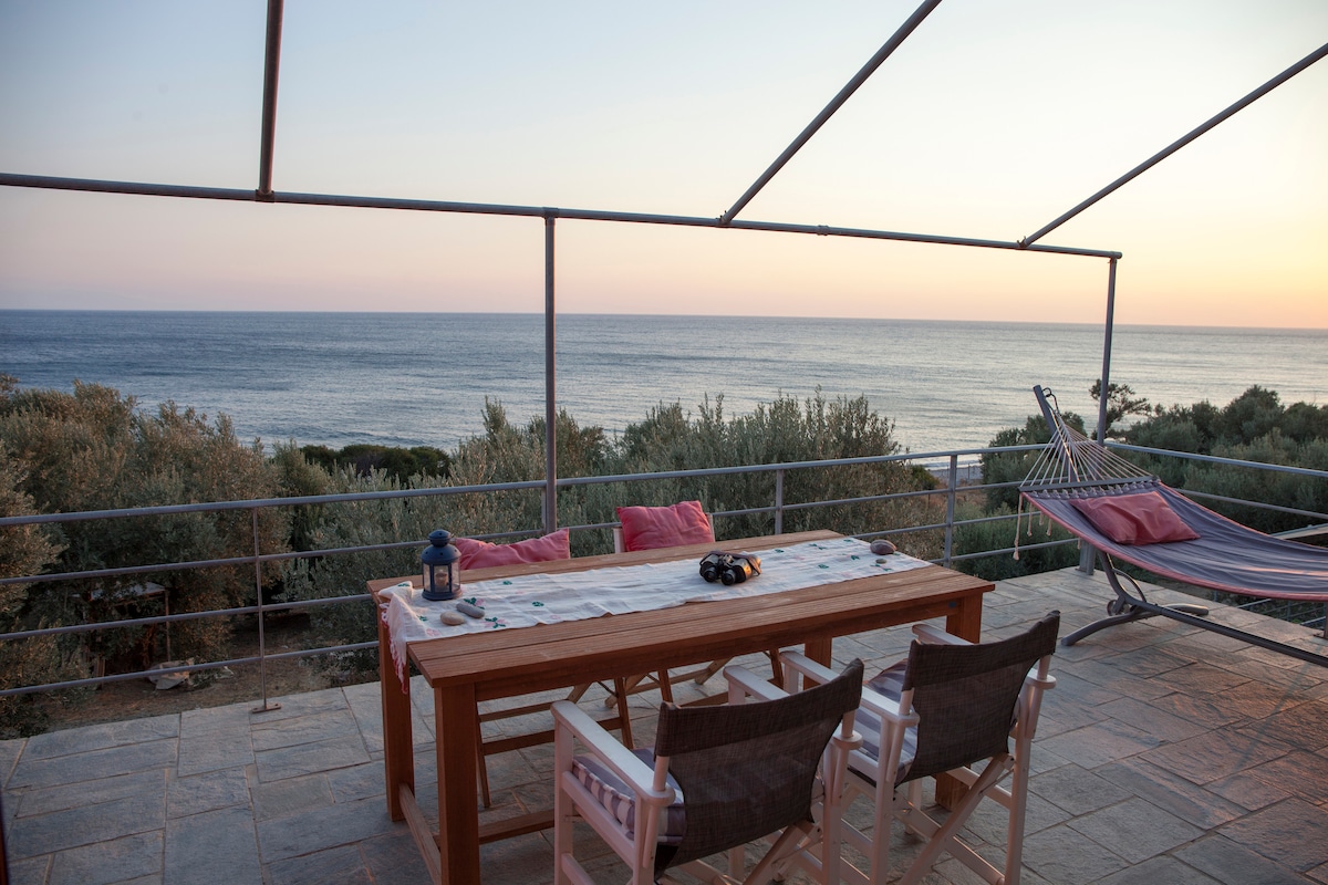 A dining area is set on a spacious veranda, featuring a wooden table with two chairs and a hammock nearby. The scene overlooks the Aegean Sea, showcasing soft hues of sunset blending into the horizon. Olive trees are visible in the background, enhancing the natural setting.
