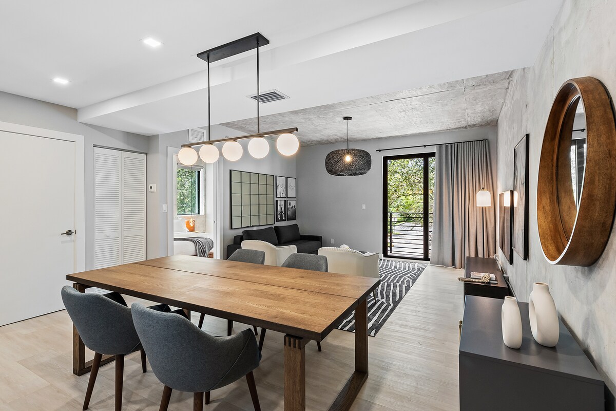 A spacious dining area features a wooden table surrounded by four upholstered chairs. A modern light fixture hangs overhead. The adjacent living space showcases a black sofa and natural light flows in through large windows, framed by curtains, enhancing the open layout.