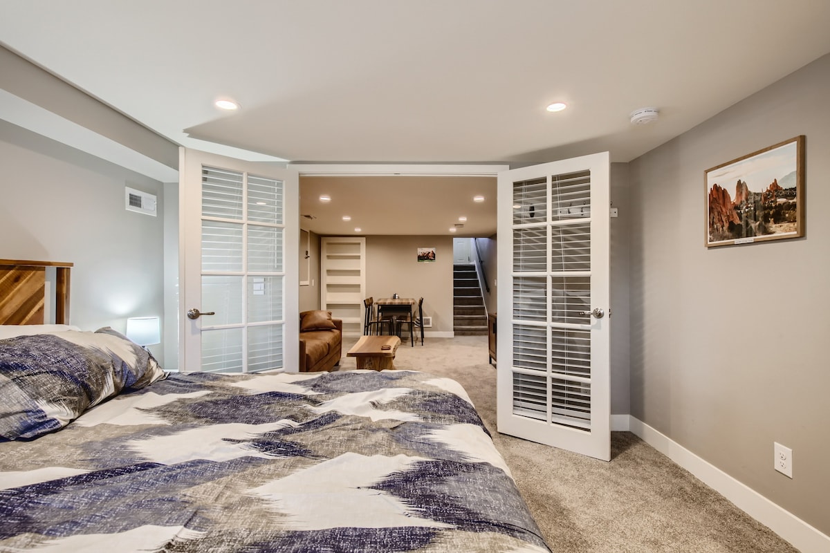 A spacious bedroom is highlighted by soft lighting and a patterned bedding set. French doors lead to a cozy living area in the background, which includes a dining table and steps leading to additional spaces within the apartment.