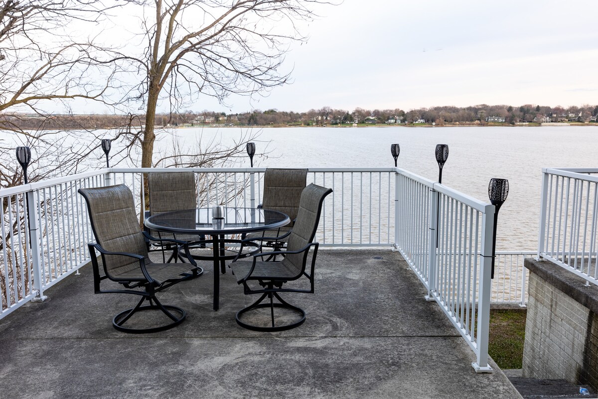 A patio area features a round table surrounded by four swivel chairs, positioned beside a railing overlooking the water. Lanterns line the edges, offering illumination. The view encompasses a broad expanse of water with trees lining the distant shore.