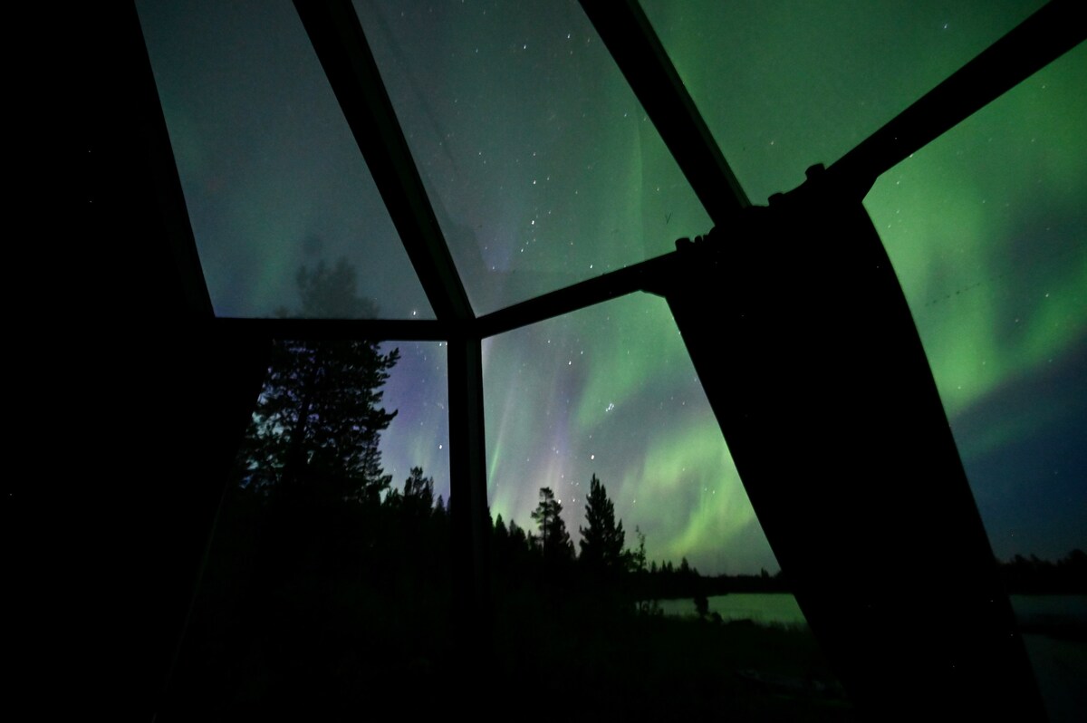 A panoramic view of the night sky is captured through large glass panels, showcasing vibrant auroras in shades of green and purple. Silhouettes of trees line the horizon, and the calm surface of the lake reflects the captivating light display.