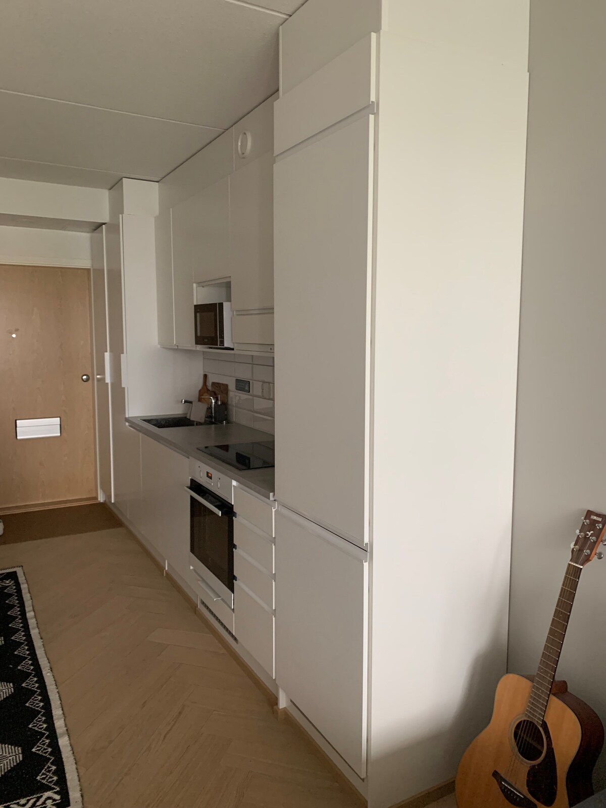 A well-organized kitchen area features modern white cabinetry and a built-in oven. A smooth countertop hosts essential appliances, including a stovetop and a microwave. Natural light illuminates the space, which includes light wood flooring and a cozy area rug. A guitar leans against the wall.