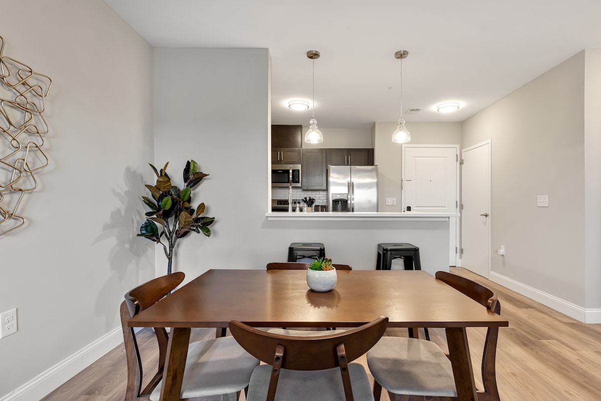 A dining area is presented with a wooden table surrounded by four chairs. A small plant is centered on the table. In the background, kitchen appliances and cabinets are visible, complemented by overhead pendant lighting and a decorative wall accent.