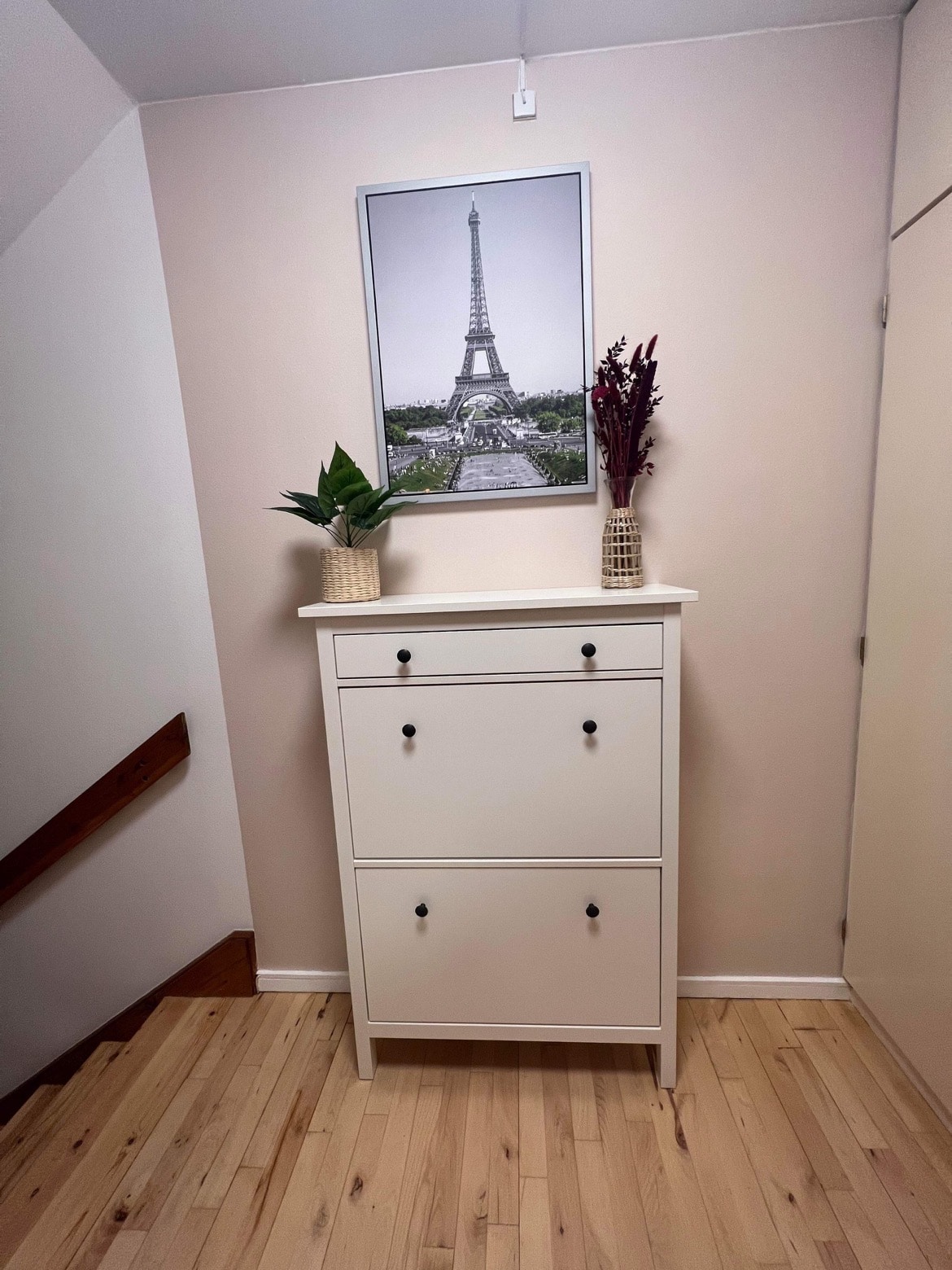A white cabinet with two drawers and two doors is positioned against a soft beige wall. Above it, a framed photograph of the Eiffel Tower is displayed. Two potted plants, featuring decorative vases, add a touch of greenery to the space.