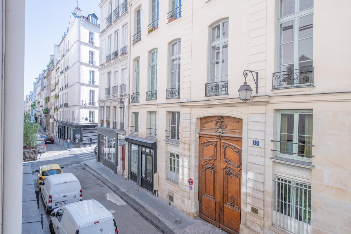 A view of a quiet street scene in Le Marais is presented, showcasing classic Parisian architecture. Elegant buildings with balconies line the street. A large wooden door is visible, along with parked vehicles and a glimpse of greenery in the background.