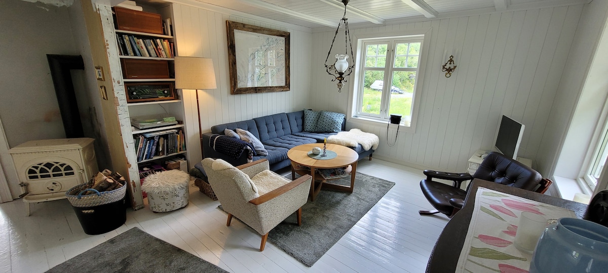 A cozy living area features a blue sectional sofa alongside a round wooden coffee table, with a plush rug beneath. Natural light enters through the windows, illuminating a white-shiplap wall adorned with framed art. A combination of chairs adds to the inviting space.