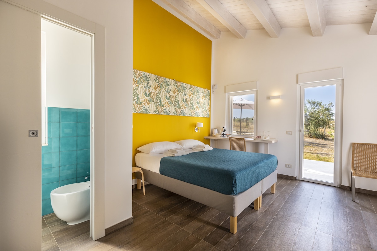 A spacious room featuring a bed with a teal blanket, complemented by a vibrant yellow accent wall. Natural light enters through two windows, illuminating a small table and chairs. An en-suite bathroom is visible through an open door, and the outdoors is accessible via a door opposite the bed.