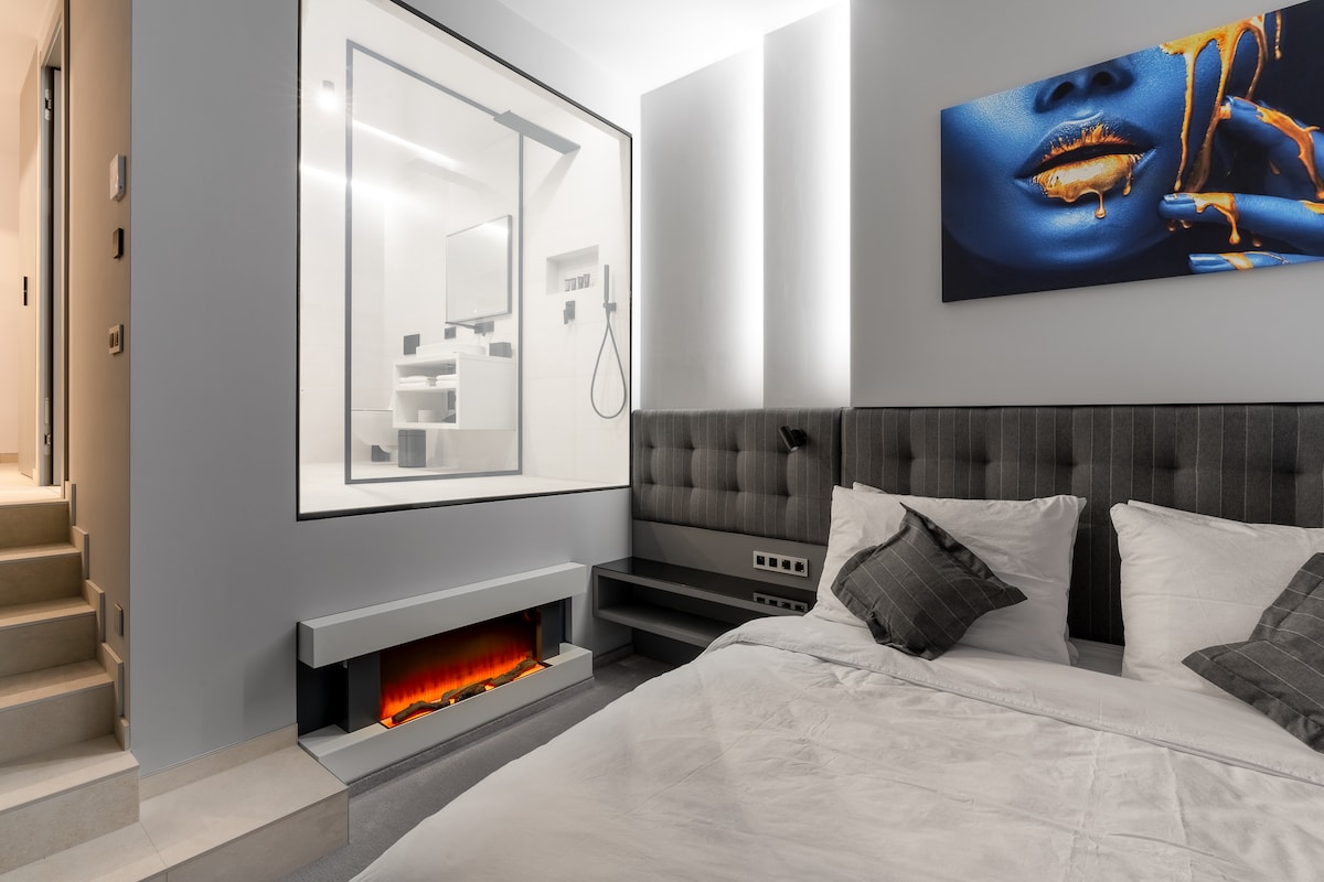 A modern bedroom features a spacious bed with plush pillows and a soft gray blanket. A sleek fireplace adds warmth, while a glass-enclosed bathroom is visible in the background, showcasing contemporary fixtures and a minimalist design. A striking piece of wall art enhances the ambiance.
