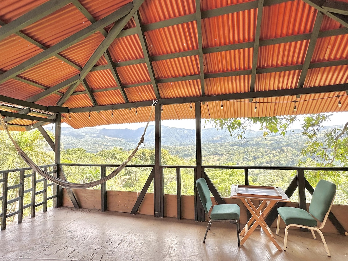 An open-air terrace features a wooden floor and a sloped roof with bright orange corrugated panels. A hammock is suspended from the beams, while a small table and two green chairs are positioned to take in the expansive mountain view.