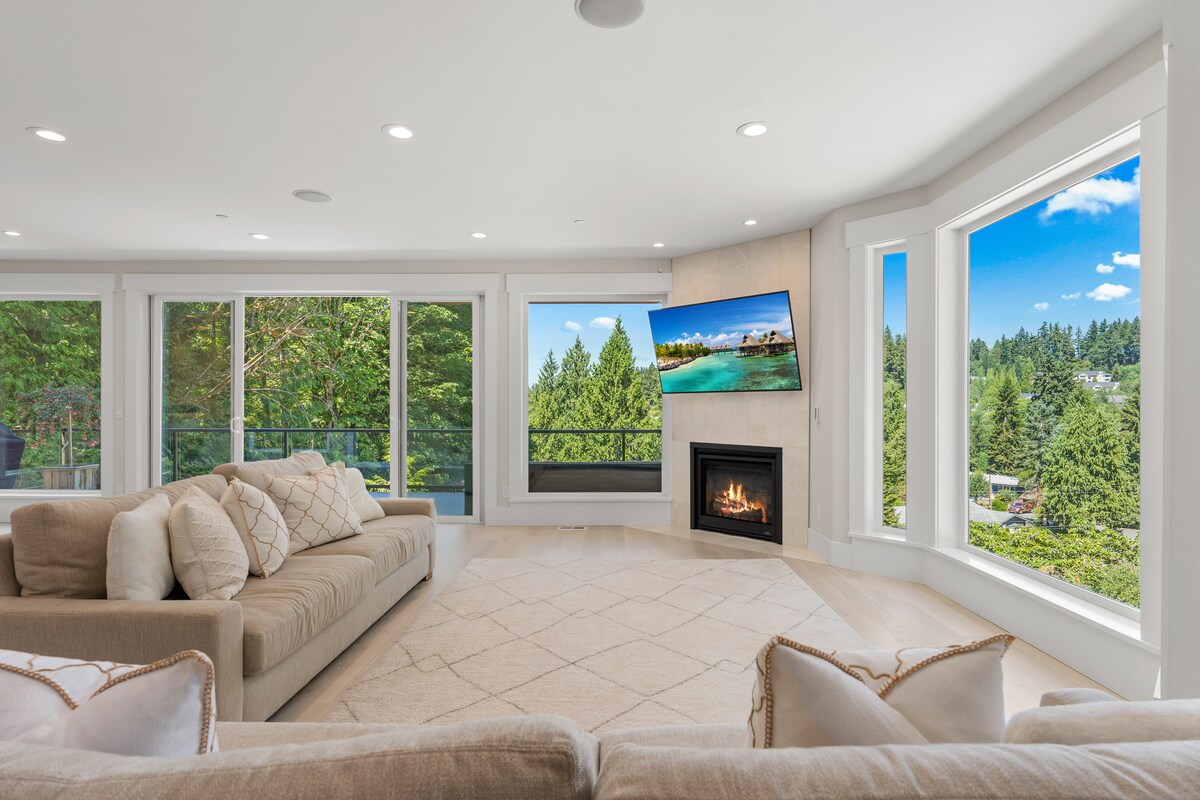 A light-filled living area is framed by large windows, offering views of the surrounding greenery. A comfortable sectional sofa sits on a neutral rug, complemented by a mounted television and a modern fireplace, creating a welcoming space for relaxation.