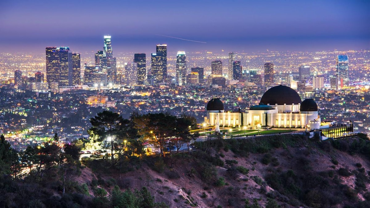 A panoramic view showcases the Los Angeles skyline at dusk, with city lights twinkling in the background. The Griffith Observatory is prominently featured in the foreground, characterized by its distinctive domes and sweeping green landscape.