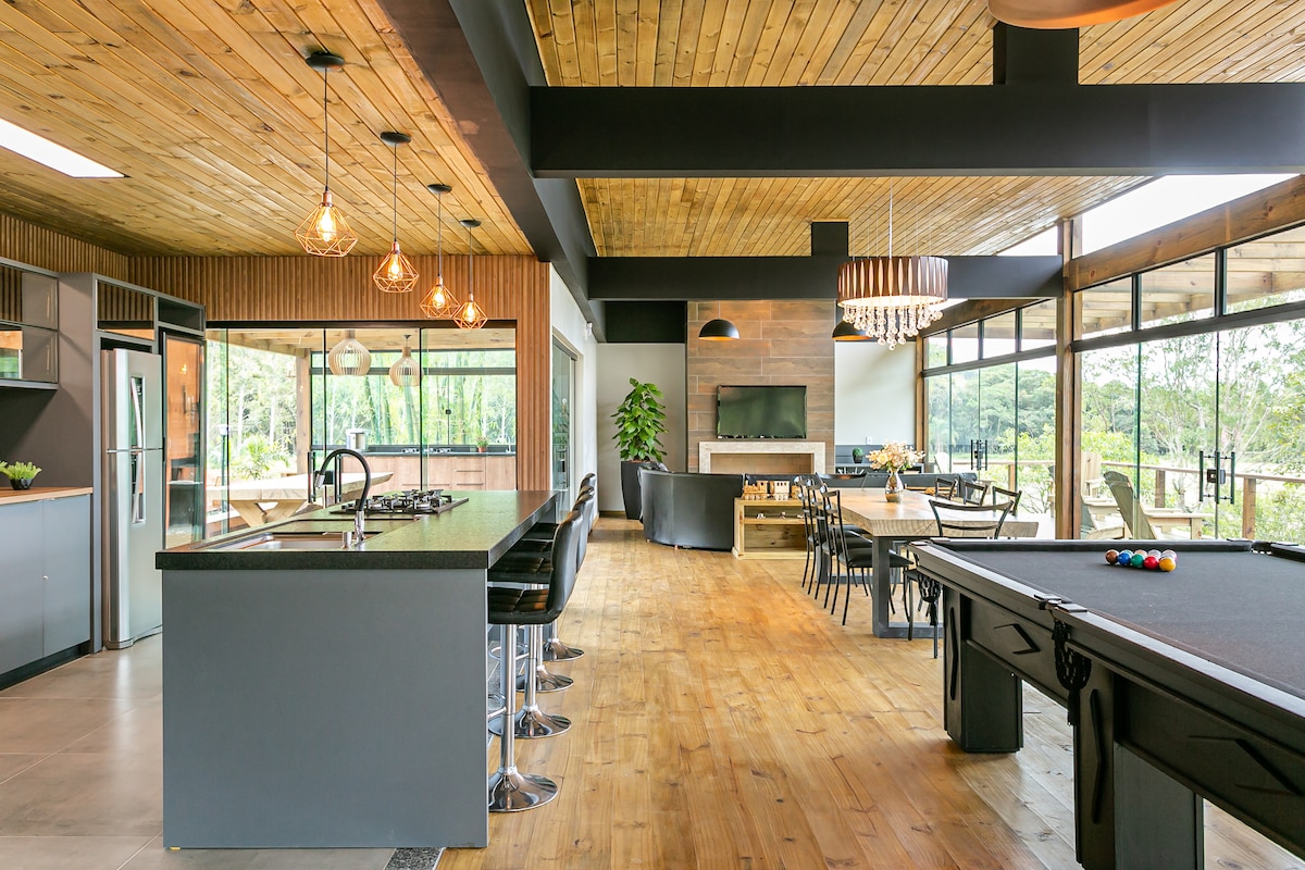 An open-plan dining and kitchen area features a large island with bar seating and modern appliances. Natural light floods through expansive windows, highlighting wooden accents throughout the space. A billiards table is positioned in the corner, providing space for entertainment while overlooking the surrounding nature.