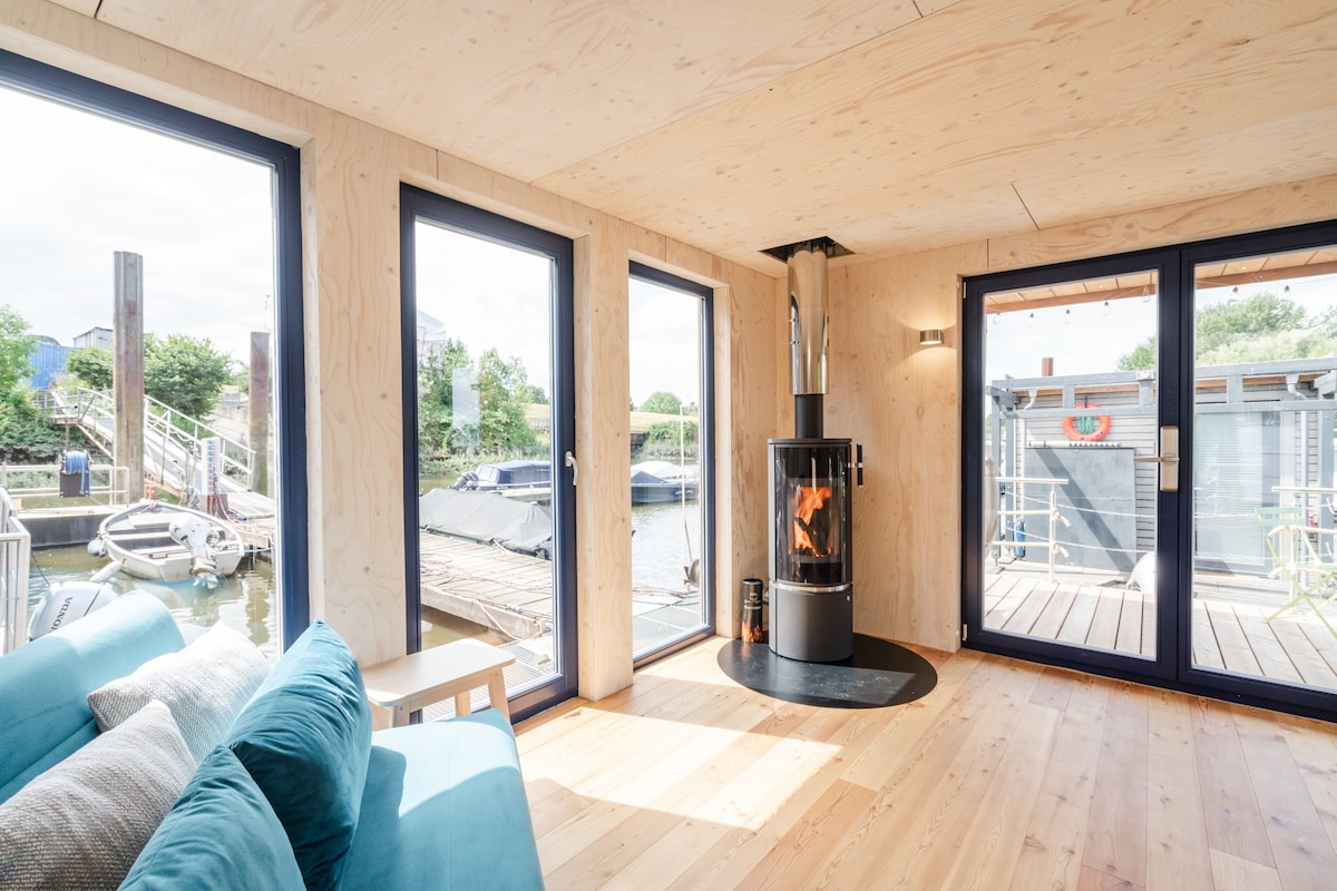 A bright living area features large windows that provide views of the surrounding water and dock. A cylindrical wood stove serves as a focal point, while a cozy sofa invites relaxation. Light wood paneling and flooring create a warm, inviting environment.