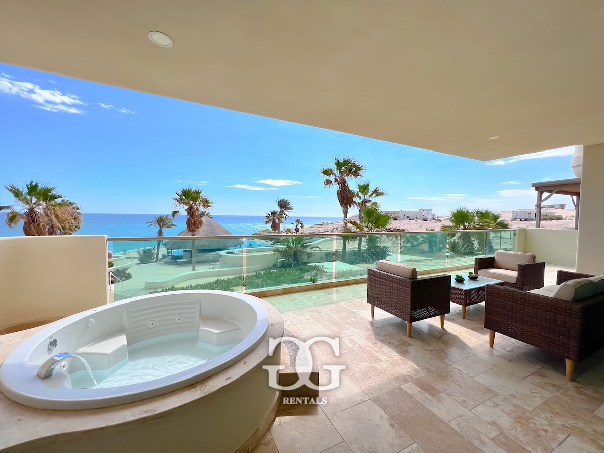 A relaxing outdoor space features a private hot tub adjacent to two comfortable chairs with matching side tables. Ocean views are showcased through large glass panels, while palm trees gently sway in the breeze under a clear blue sky.