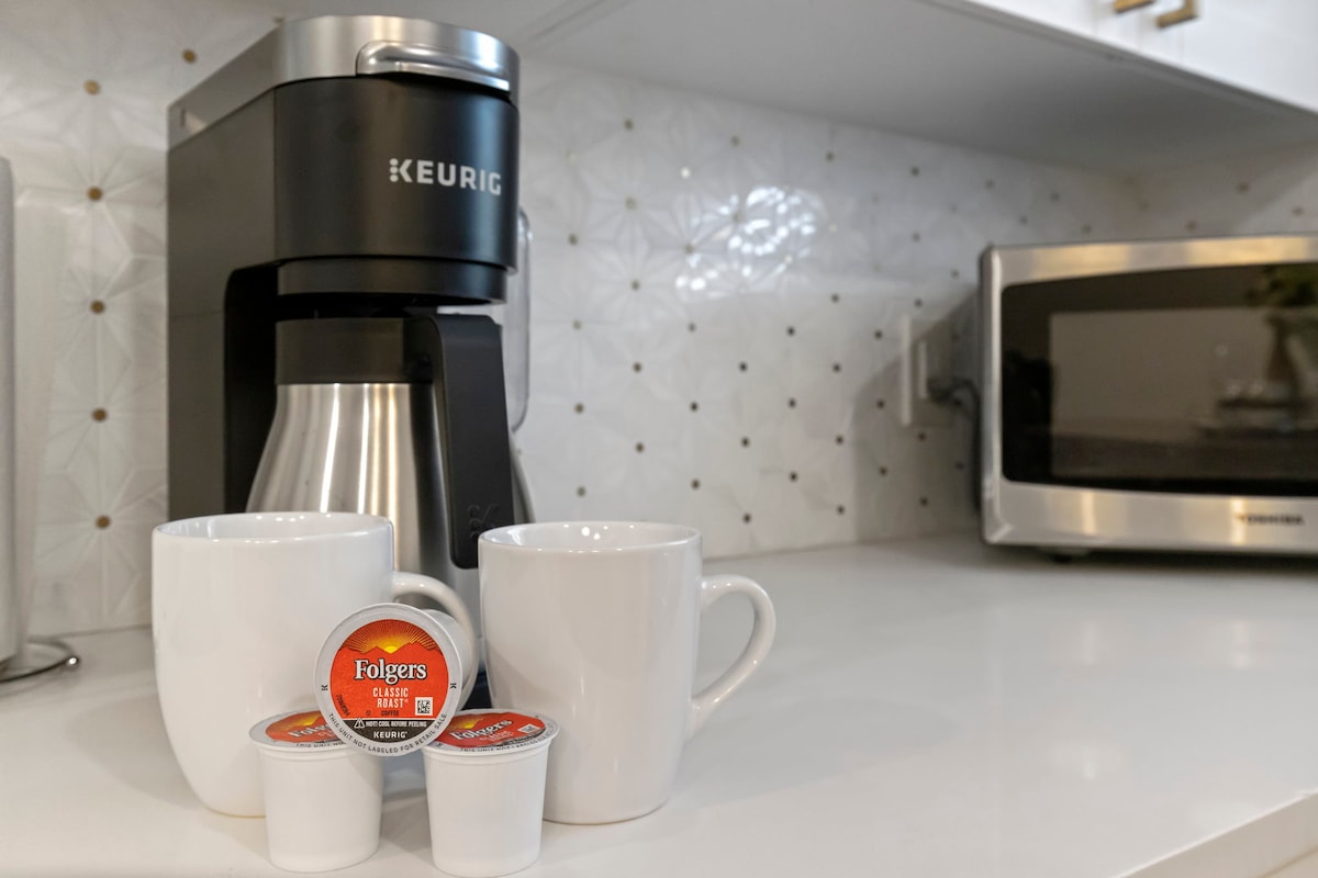 A coffee station is arranged on a clean countertop, featuring a Keurig coffee maker and two white mugs. Several coffee pods are neatly placed in front of the mugs, with a microwave visible in the background against a decorative tile wall.
