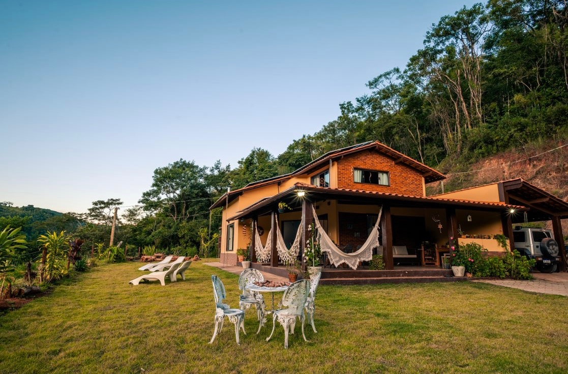 A two-story house is surrounded by lush greenery, with a spacious lawn in the foreground. Outdoor seating, including white chairs and a table, is positioned on a patio area. Hammocks are gracefully draped from the porch, complementing the serene mountain backdrop.