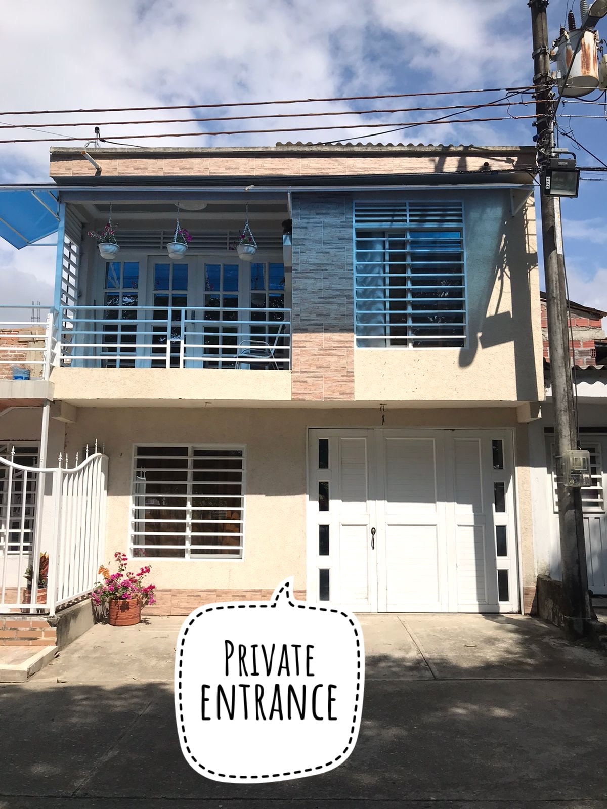 A modern exterior of a two-story apartment building is shown, featuring a light-colored façade with decorative stone accents. Large windows allow natural light to enter. The entrance is private, marked by a gated pathway and potted plants enhancing the welcoming appearance.