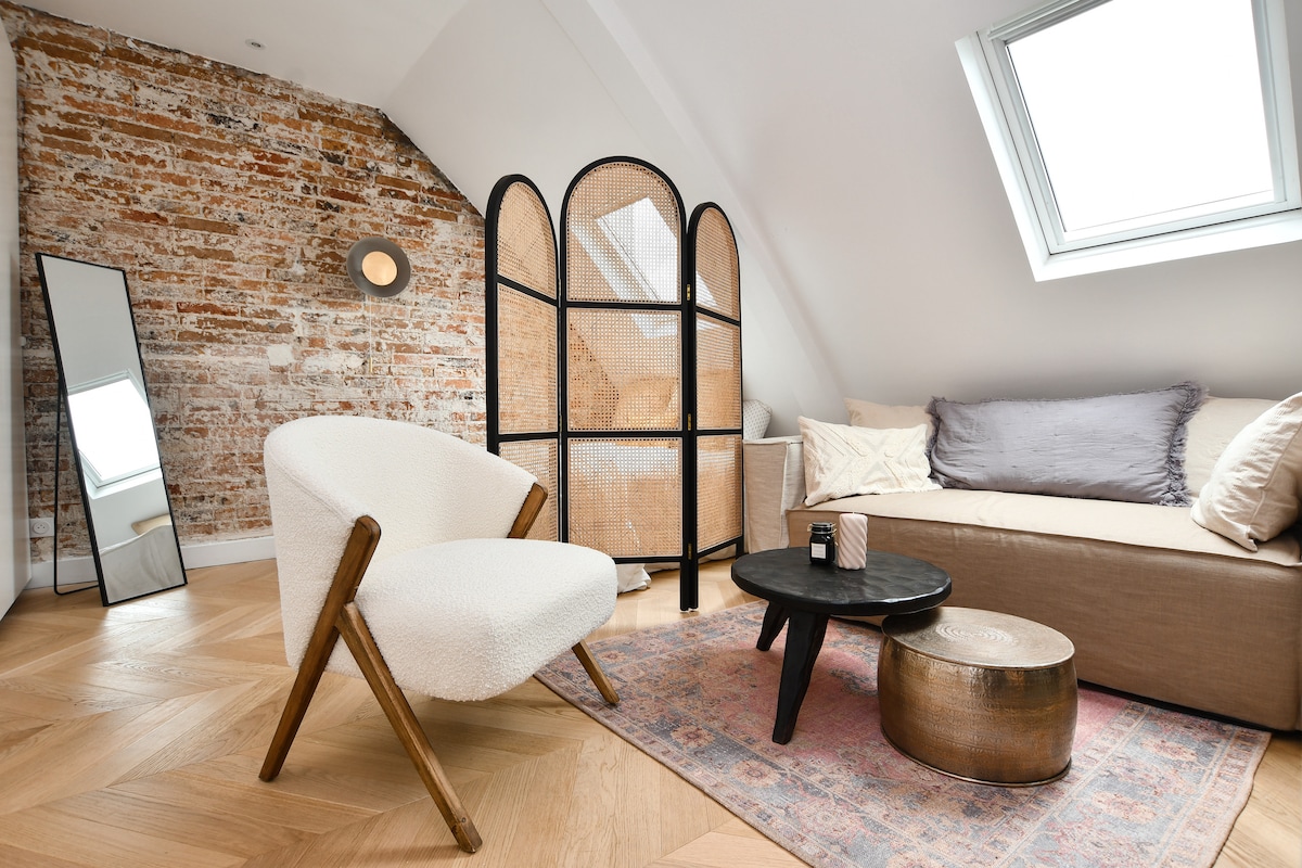 The living area features a soft armchair and a low round table on a textured rug. Exposed brick walls add character, while natural light enters through a skylight. A mirror reflects the space, contributing to the bright atmosphere. A cozy sofa is placed against the wall.