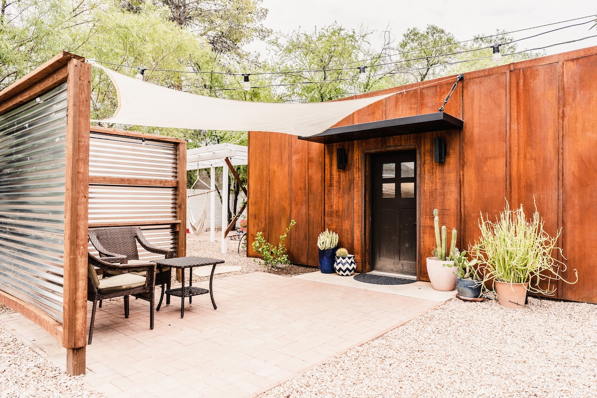 An outdoor seating area is positioned near the entrance, featuring two chairs and a small table. Surrounding greenery includes potted plants and cacti. A fabric canopy provides shade, while overhead lights create an inviting atmosphere. The rustic structure's metal and wood textures are prominently displayed.