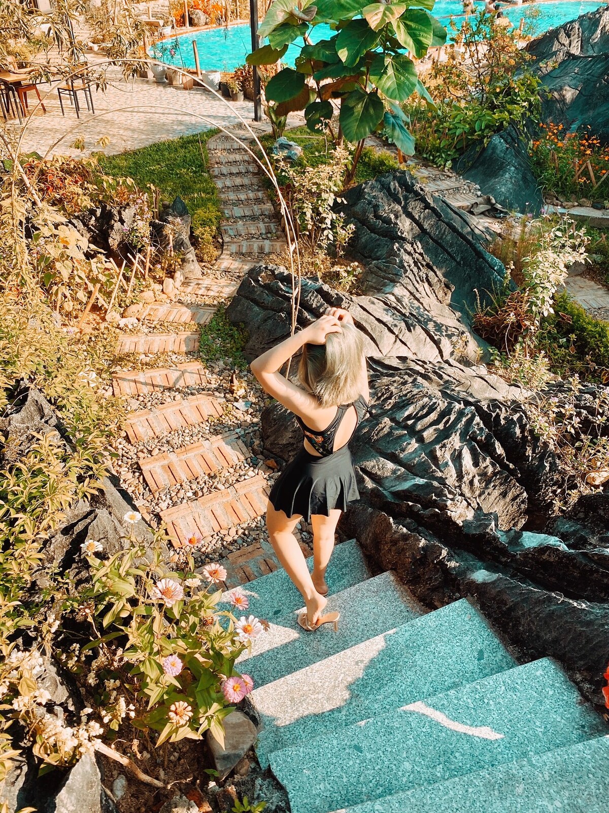 A winding stone staircase surrounded by lush greenery and colorful flowers leads through a landscaped garden. The steps, bordered by dark rocks, create a natural path to a pool in the background, providing a serene atmosphere for relaxation.