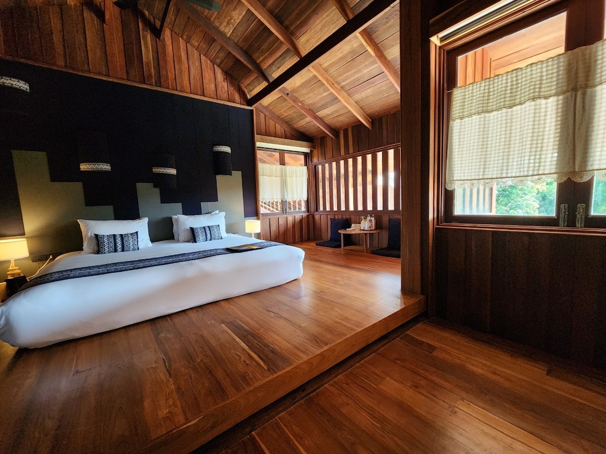 A spacious bedroom features a large bed with a minimalist design, complemented by warm wooden walls and flooring. Natural light filters through large windows with sheer curtains, illuminating a cozy seating area and a small table against the backdrop of greenery outside.