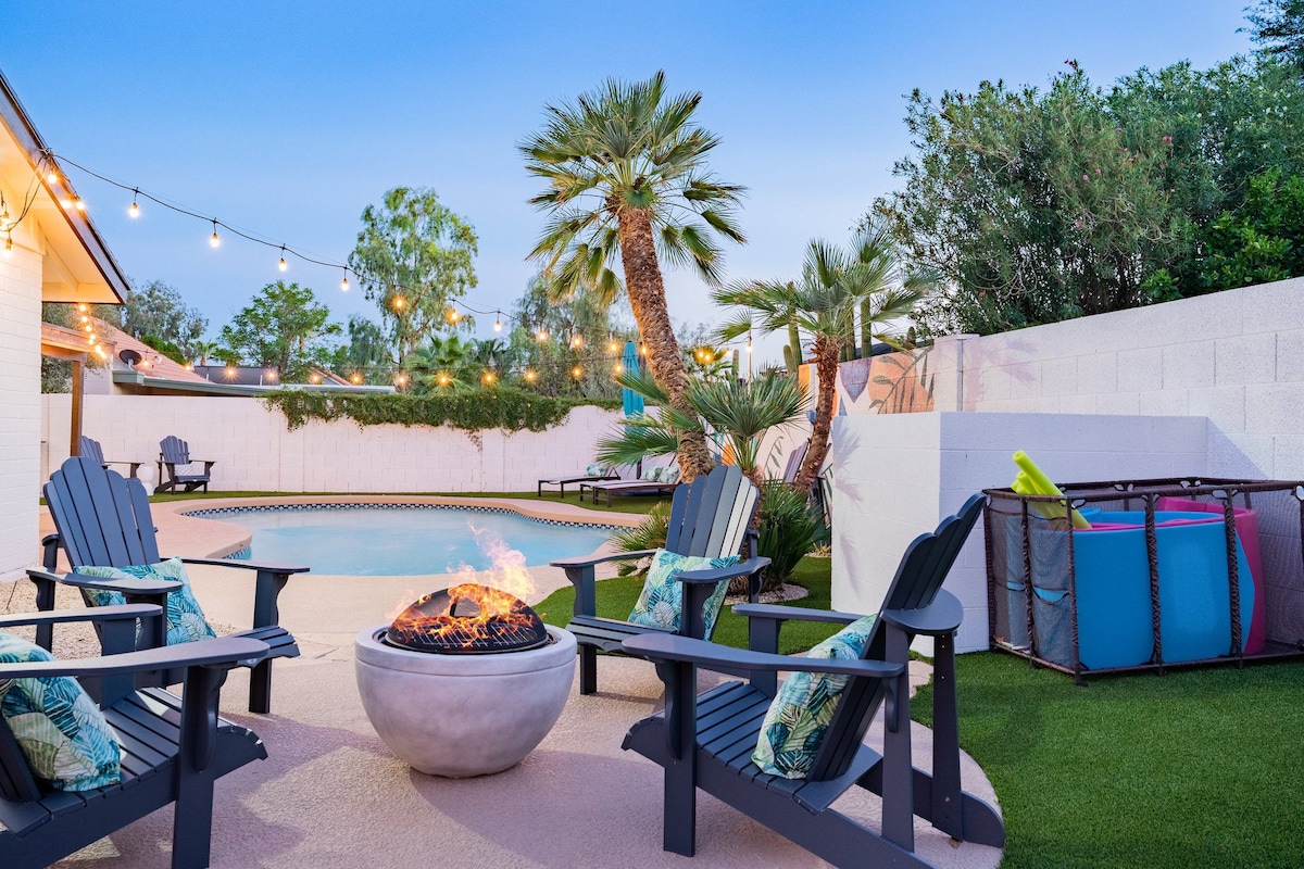 A vibrant backyard features a wood-burning fire pit surrounded by four Adirondack chairs, accented with tropical cushions. Lush palm trees line the space, and the heated pool is visible in the background, set against a well-maintained landscape. Colorful pool floats are neatly arranged nearby.