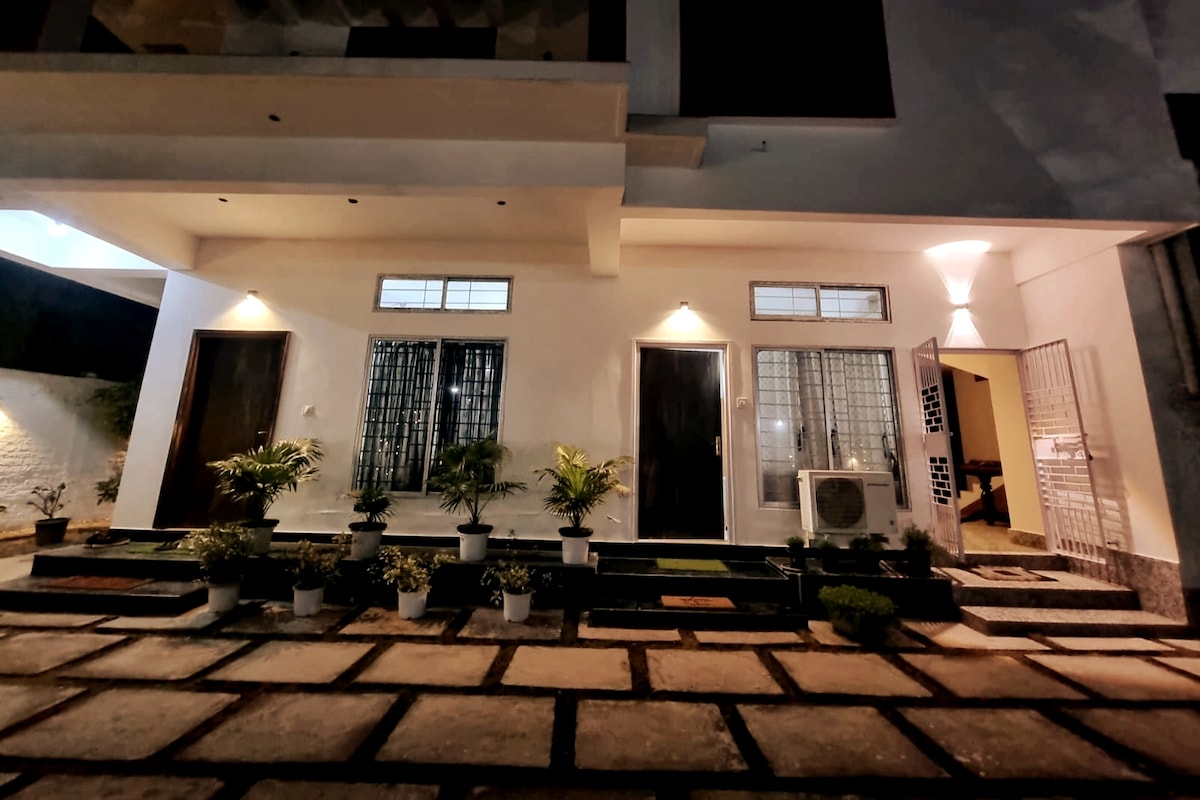 The exterior entrance features a well-lit walkway, showcasing a modern facade with three doors. Lush green plants in white pots line the steps, and an air conditioning unit is positioned to the side, enhancing the inviting atmosphere of the entrance.