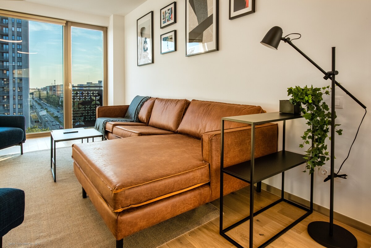 A modern living area is featured, showcasing a spacious brown sectional sofa and a sleek black side table. A floor lamp provides soft lighting, while artwork decorates the walls. Large windows offer views of the surrounding cityscape, enhancing the bright ambiance of the space.