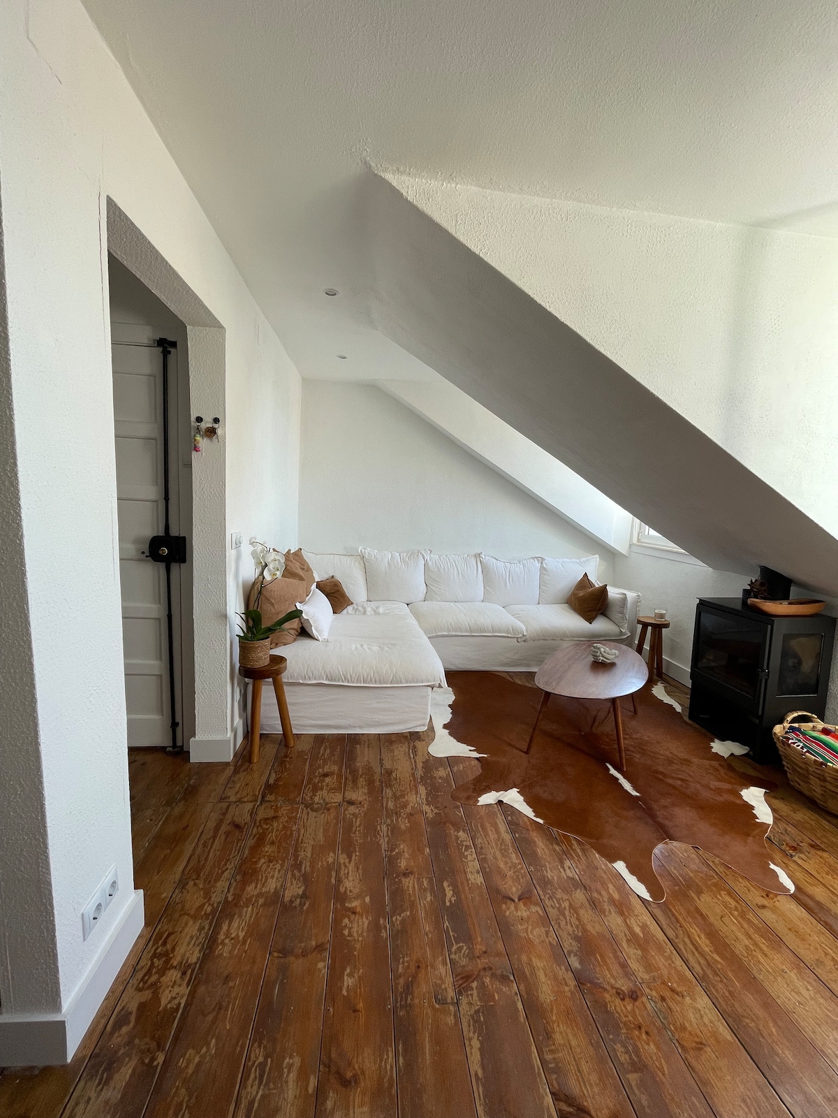 A bright living space is highlighted by sloped ceilings and features a white sofa alongside a small round table. A decorative cowhide rug adds warmth to the wooden floor, while a black stove creates a cozy setting. Natural light fills the room through nearby windows.