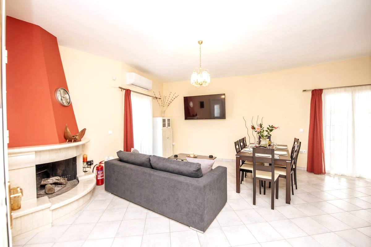 A spacious living area features a comfortable gray sofa arranged near a fireplace and a wall-mounted television. A dining table for eight is positioned nearby, with large windows allowing natural light to fill the room. Curtains frame the windows, enhancing the ambiance.