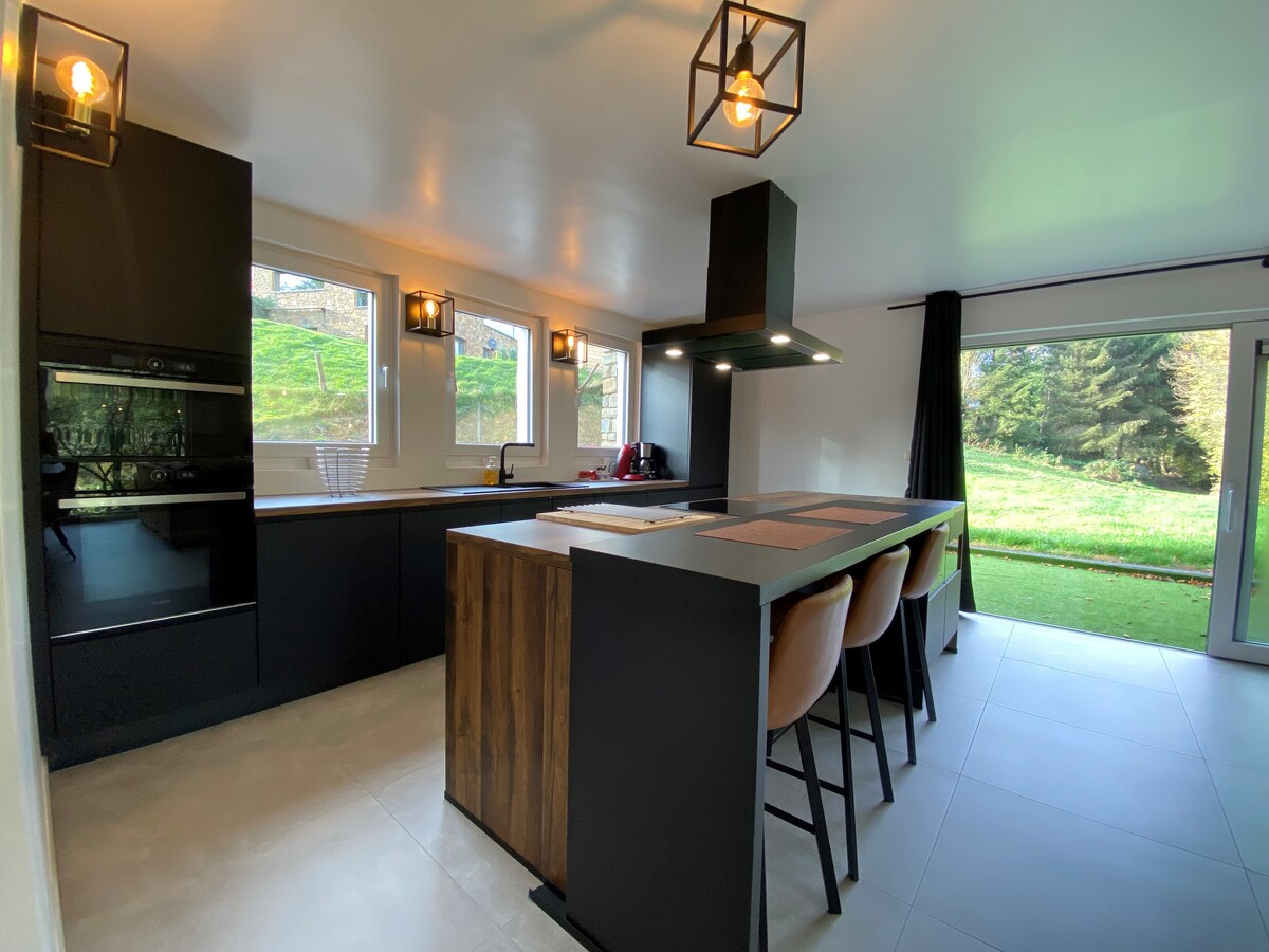A modern kitchen equipped with sleek black cabinetry and an island featuring wooden accents. Three bar stools are positioned at the island. Large windows allow natural light, with a view of greenery visible through glass doors.