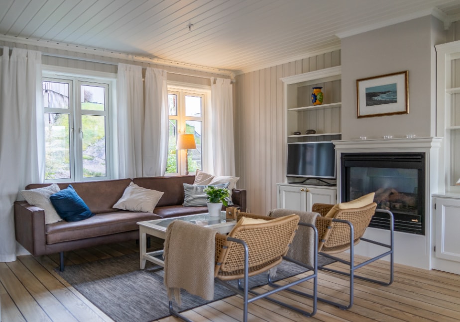A cozy living area features light-colored walls and wooden flooring. A brown sectional sofa is complemented by throw pillows in various shades. A coffee table sits in the center, surrounded by two wicker chairs. Natural light streams through the windows, and a fireplace is positioned against one wall.