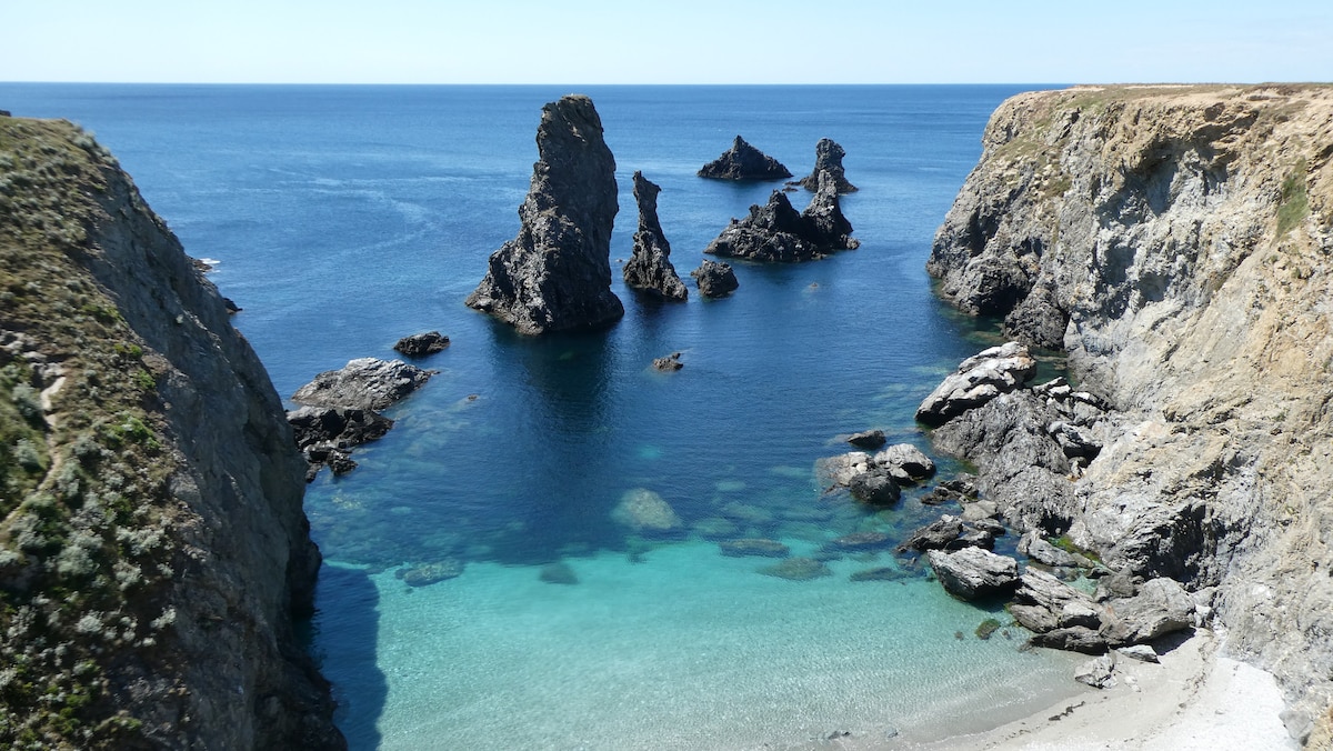 A serene coastal landscape is presented, featuring rugged rock formations rising from calm turquoise waters. The shoreline is adorned with soft sandy areas, while the surrounding cliffs are covered in vegetation, offering a natural and unspoiled view of the Brittany coastline.