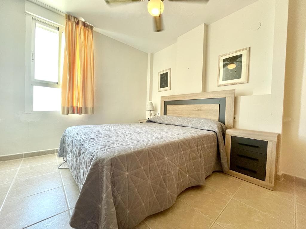 A sleeping area features a spacious bed covered with a textured grey quilt. Nightstands are positioned on either side of the bed, complementing the room's minimalistic decor. Natural light filters through a large window with sheer curtains, enhancing the soft ambiance.