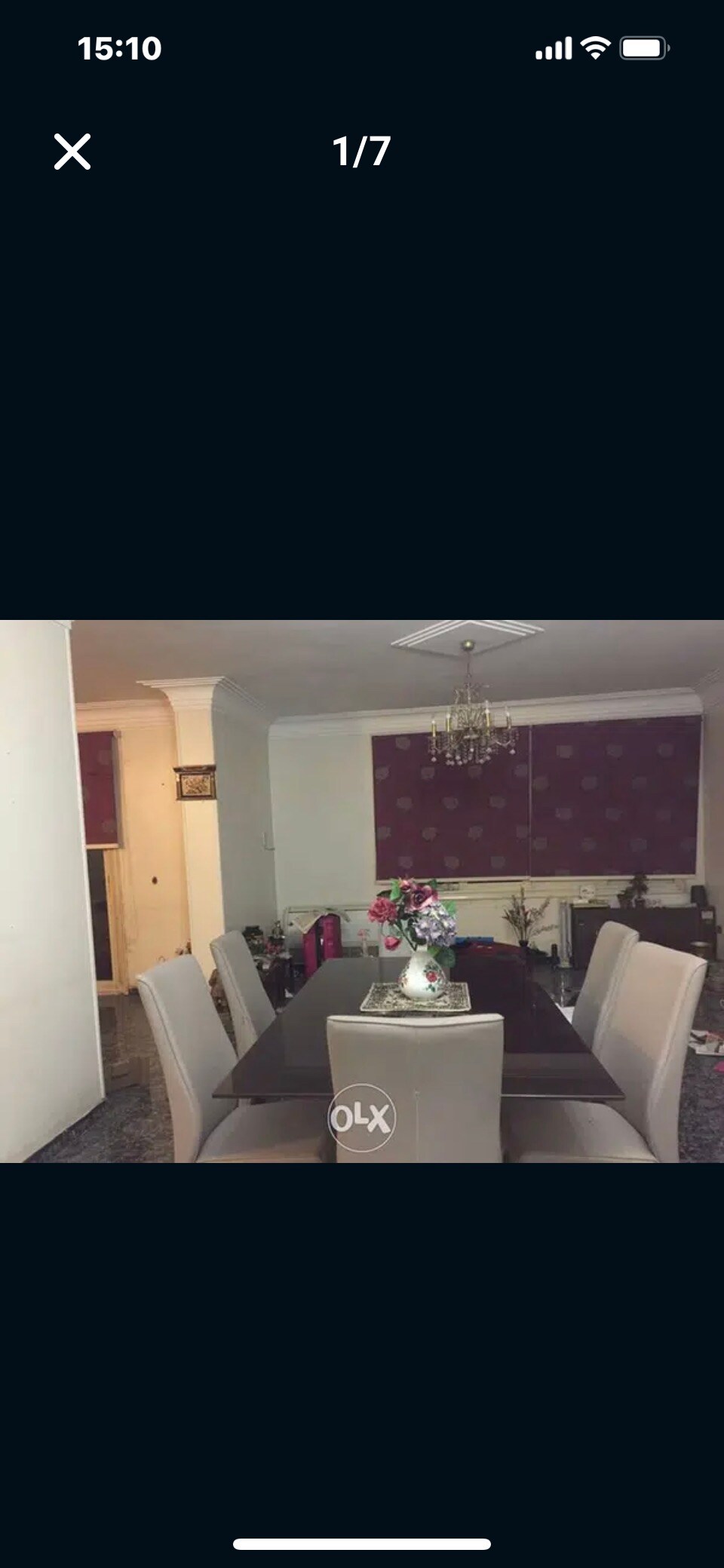 A dining area is shown with a long, dark table surrounded by six padded chairs. A centerpiece of flowers is displayed on the table. A chandelier hangs from the ceiling, complementing the patterned wall behind, while a cozy atmosphere is enhanced by soft lighting.