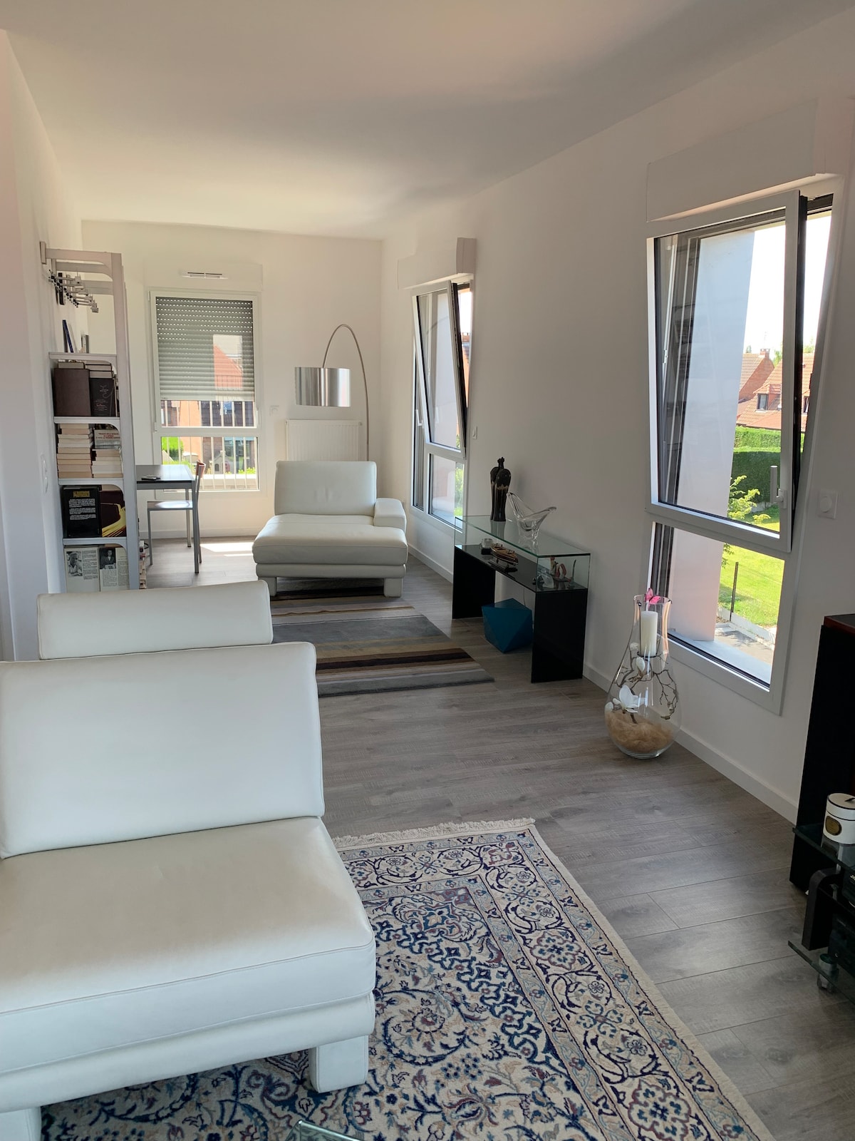 A spacious living area features light-colored furniture and large windows that allow ample natural light. A soft area rug complements the seating, while decorative items are displayed on a sleek glass table. The view beyond includes greenery, creating a connection with the outdoors.