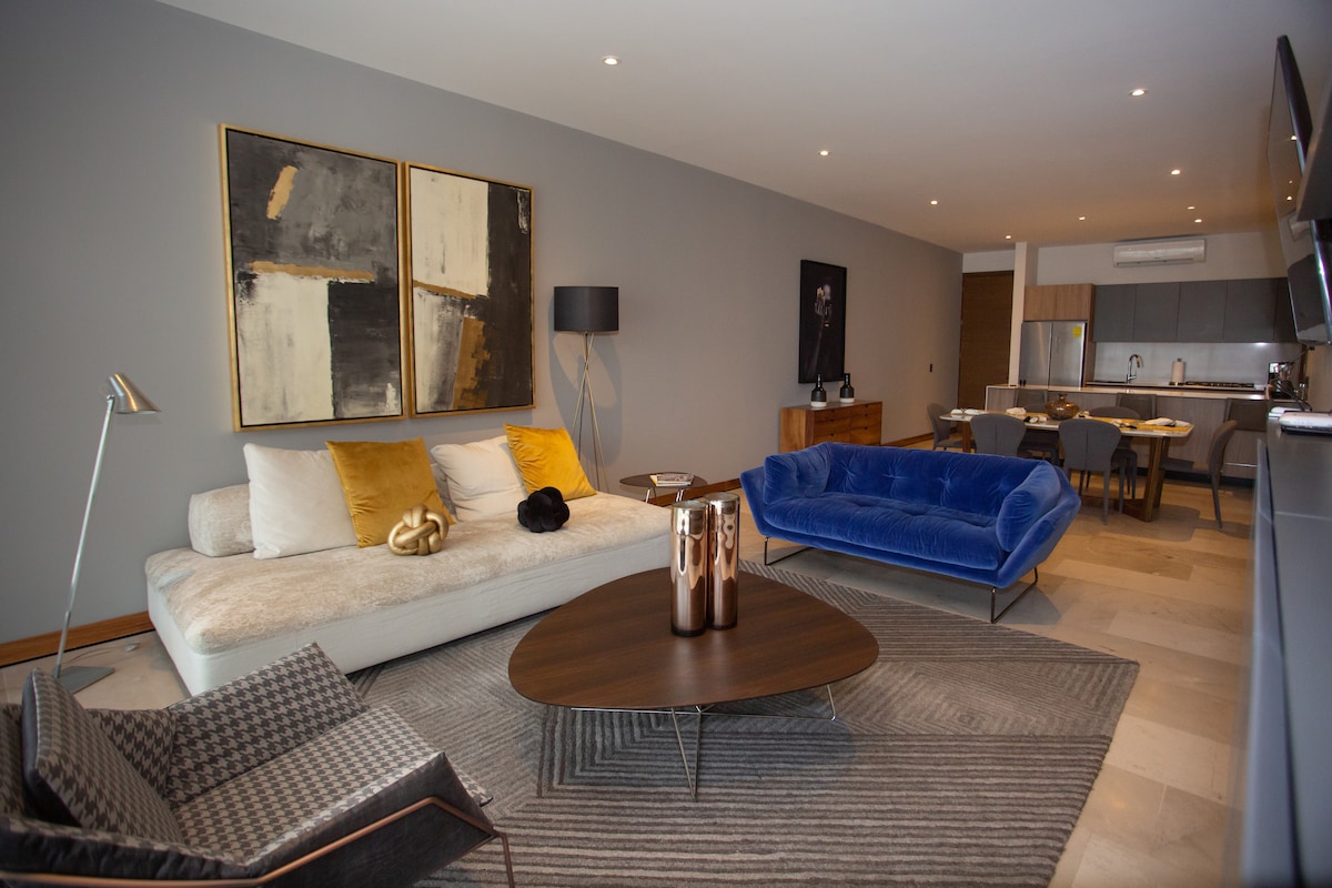 A modern living area features a plush sectional sofa complemented by a deep blue velvet couch. Abstract art adorns the walls, while a sleek coffee table stands at the center. Soft lighting from a floor lamp enhances the inviting ambience.