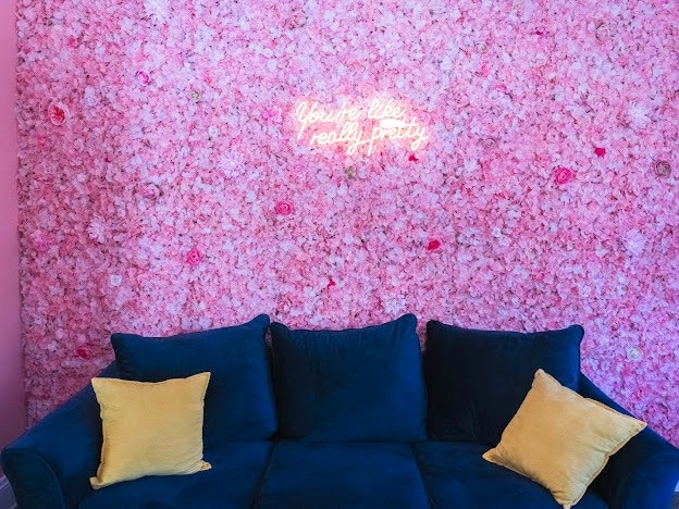 A soft blue couch is positioned in front of a vibrant pink floral wall, adorned with decorative flowers. A neon sign displaying the phrase 'You're like, really pretty' adds a playful touch to the space. Two yellow cushions are placed on the couch for comfort.