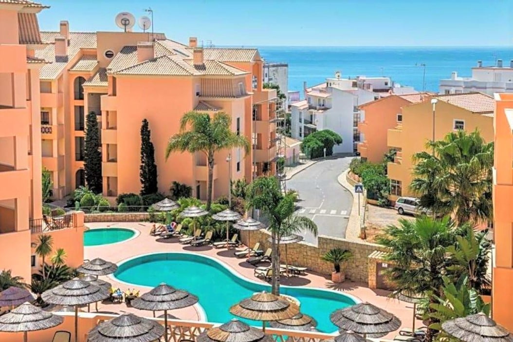 A serene view of the outdoor pool area is featured, surrounded by lounge chairs and umbrellas. The vibrant blue of the pool is contrasted by the soft hues of the neighboring buildings and the lush greenery. The distant sea complements the scenic setting.
