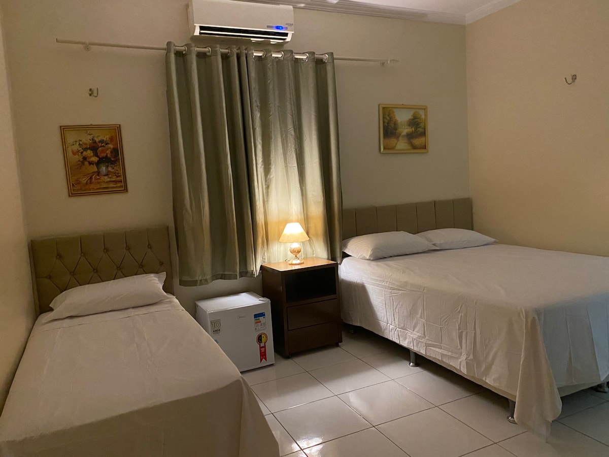 A comfortable bedroom is presented with two beds, one double and one single. Light-colored linens are neatly arranged, and a bedside table with a lamp provides soft lighting. A small refrigerator is seen in the corner, and subtle artwork decorates the walls.