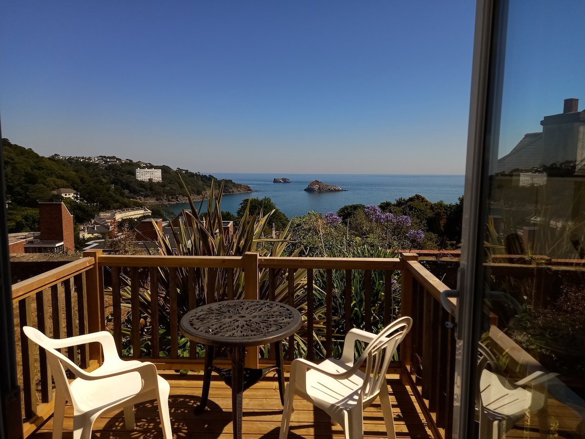 The exclusive decking area features a round table surrounded by two white chairs. A panoramic view of Thatcher's Rock and the sapphire sea is visible beyond, framed by lush greenery and vibrant flowers under a clear blue sky.