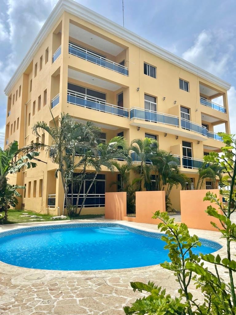 A modern four-story building showcases a vibrant yellow exterior with multiple balconies. Lush greenery surrounds a private swimming pool, which features a textured stone deck. The sky is partly cloudy, creating a bright and airy atmosphere around the property.