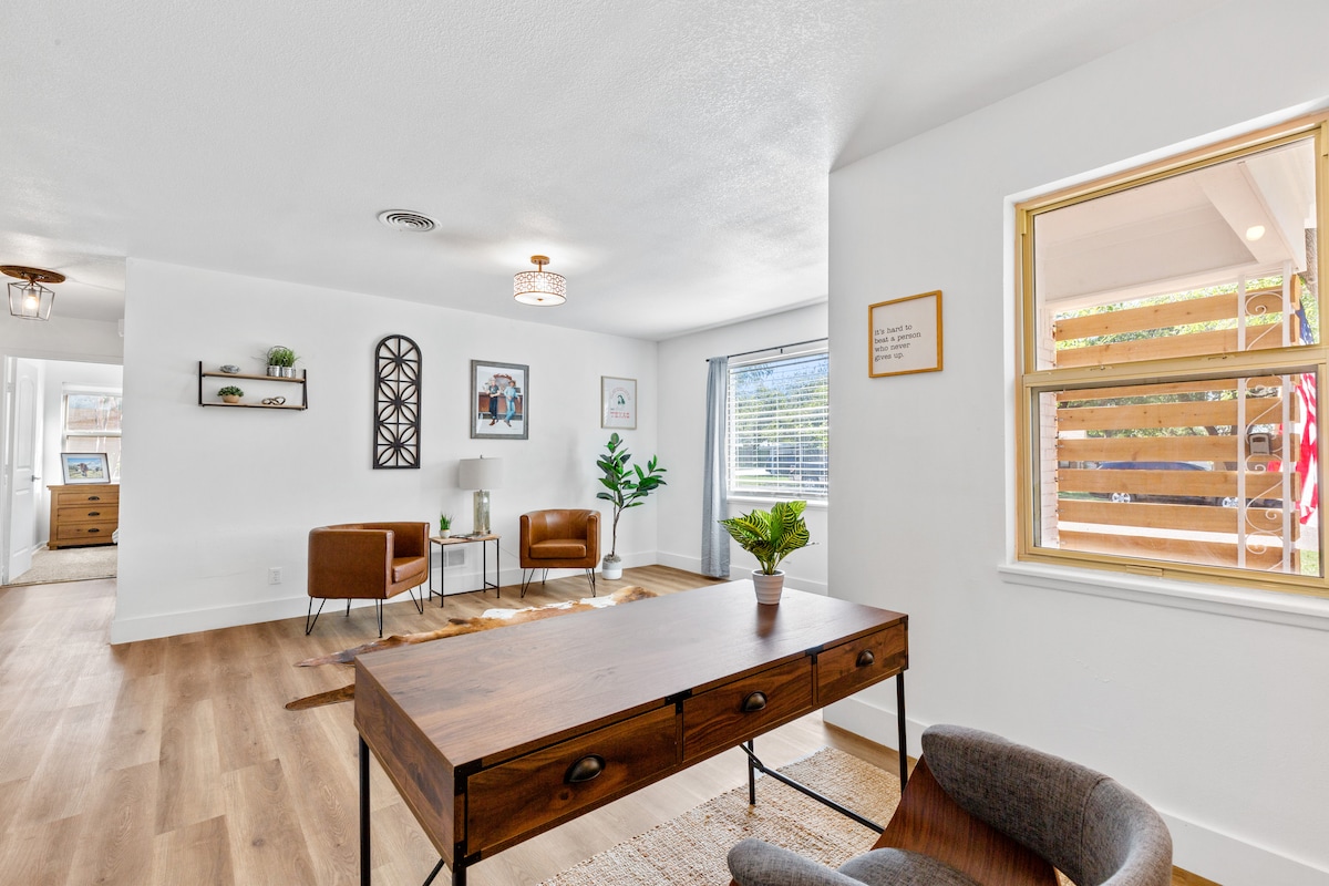 A bright living space is featured with warm wood flooring and natural light pouring in through the window. A stylish desk sits in the foreground, accompanied by an accent chair. Two leather chairs and decorative plants create a welcoming atmosphere.
