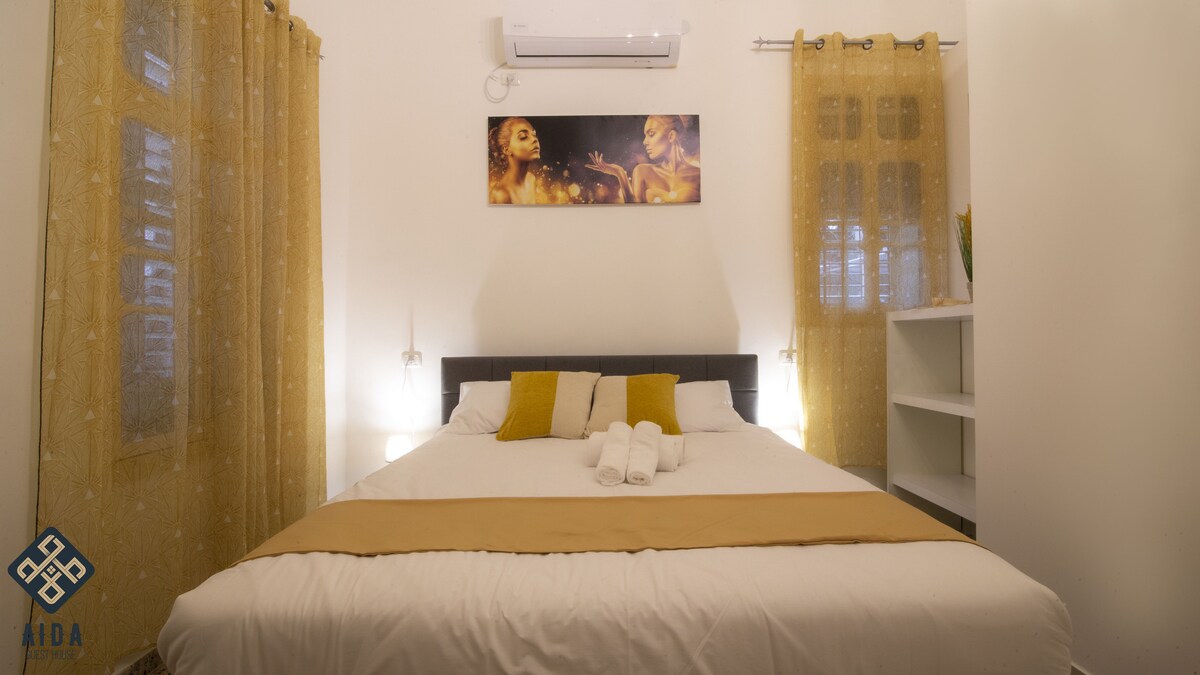 A comfortable sleeping area features a spacious bed adorned with soft linens and decorative pillows. Light-colored walls enhance the brightness of the room, while sheer curtains frame the windows. A modern air conditioning unit is positioned above, providing climate control.